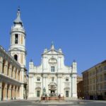 Loreto un luogo antico che parla all'anima: la facciata bianca della Basilica domina la piazza con la fontana e il campanile.