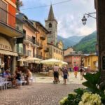 Un gioiello alpino: Limone Piemonte. Scorcio sulla piazza acciottolata con caffè all'aperto, storiche case colorate e l'imponente campanile in pietra della chiesa contro le montagne boscose, incorniciato da ortensie fiorite e lampioni stile antico.