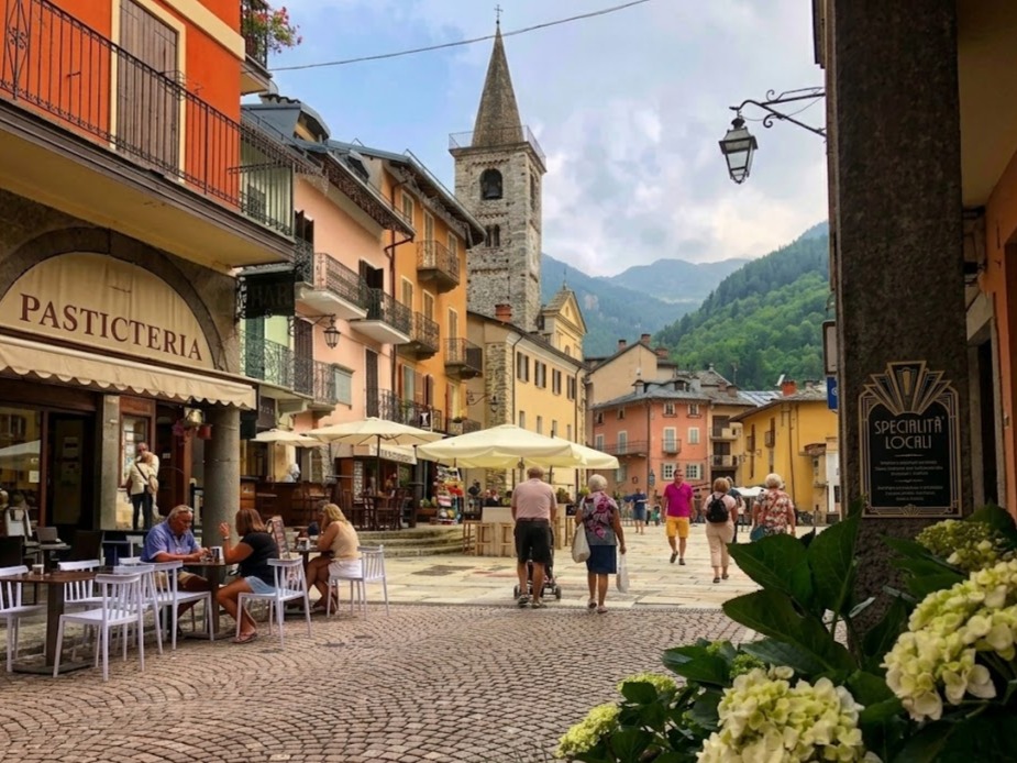 Un gioiello alpino: Limone Piemonte. Scorcio sulla piazza acciottolata con caffè all'aperto, storiche case colorate e l'imponente campanile in pietra della chiesa contro le montagne boscose, incorniciato da ortensie fiorite e lampioni stile antico.