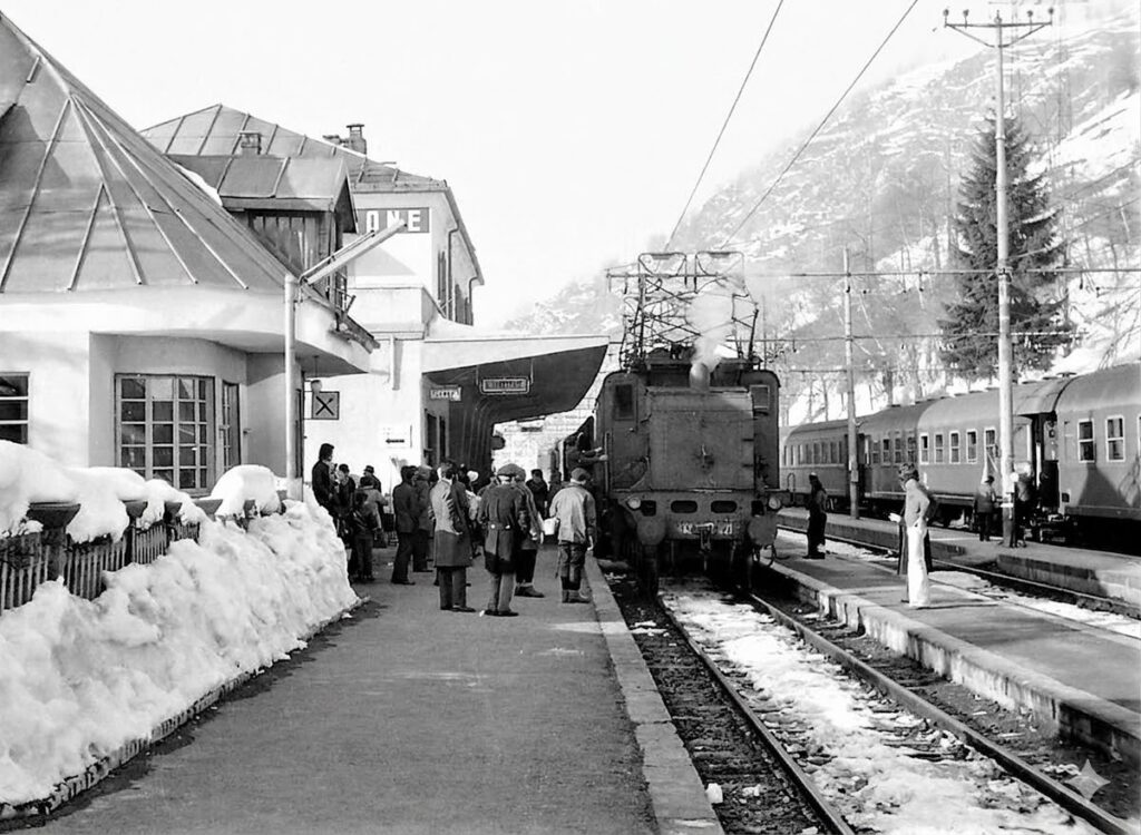 Un gioiello alpino: foto storica della stazione di Limone Piemonte innevata. Un treno è fermo sui binari, circondato da passeggeri in attesa sulla banchina e dalle montagne circostanti, catturando un momento intramontabile della sua storia.
