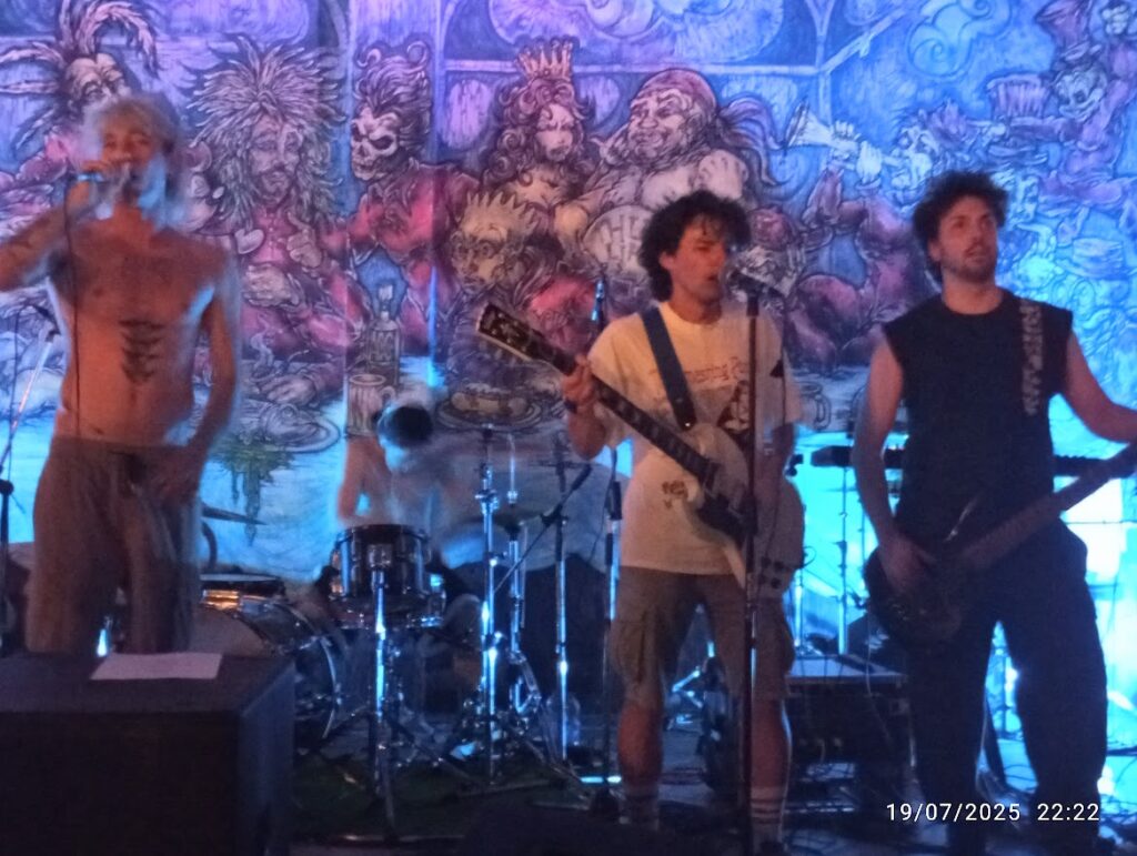 Andrea Ale Peiretti e Mane si esibiscono dal vivo con la band su un palco davanti a un murale artistico colorato