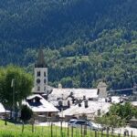 Foto: borgo alpino con chiesa e campanile, tetti in pietra, strada con auto e prati in primo piano, circondati da foreste e montagne. Cose da fare a La Salle