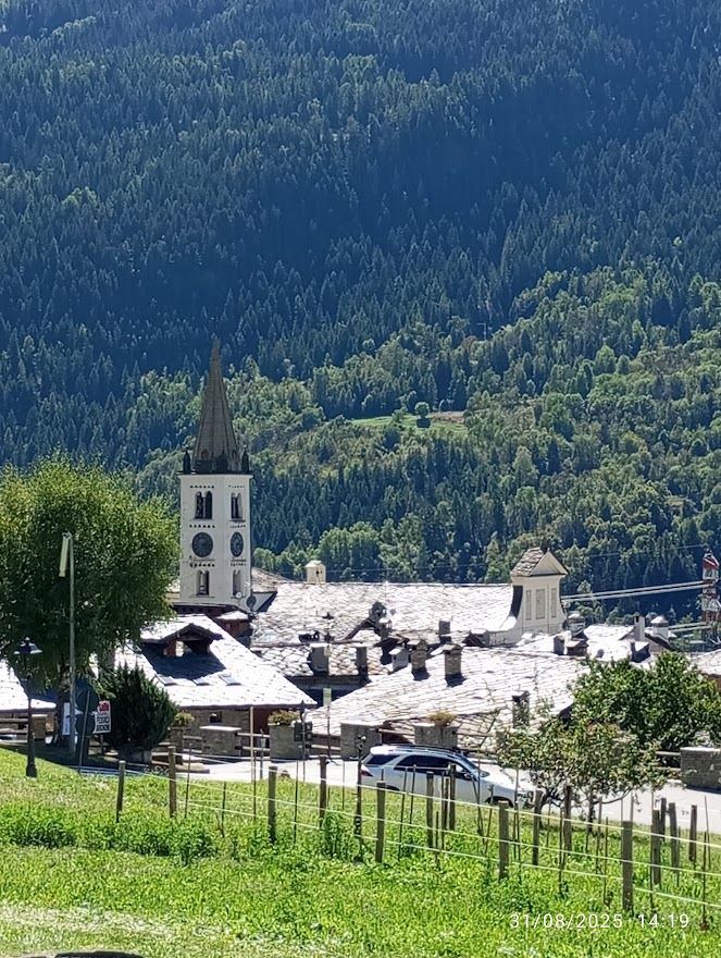 Foto: borgo alpino con chiesa e campanile, tetti in pietra, strada con auto e prati in primo piano, circondati da foreste e montagne. Cose da fare a La Salle